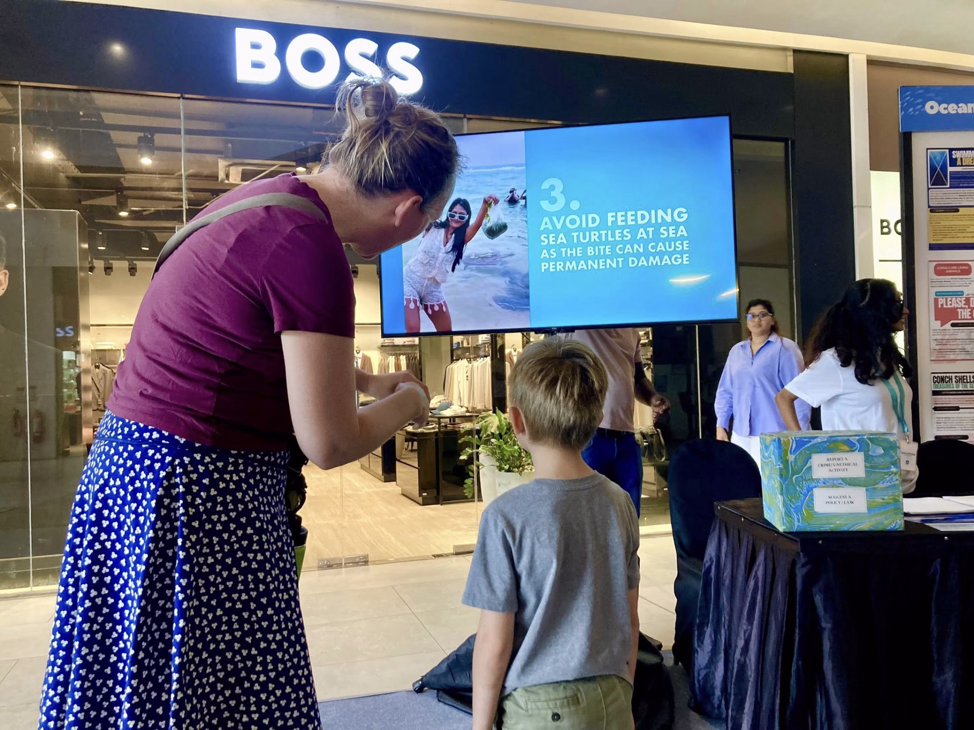 A mother and child read an educational screen advising against feeding sea turtles, inside the World Oceans Day awareness zone.